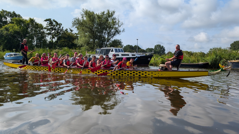 Groep mensen roeit in een gele drakenboot op een kanaal, omringd door natuur en andere boten.