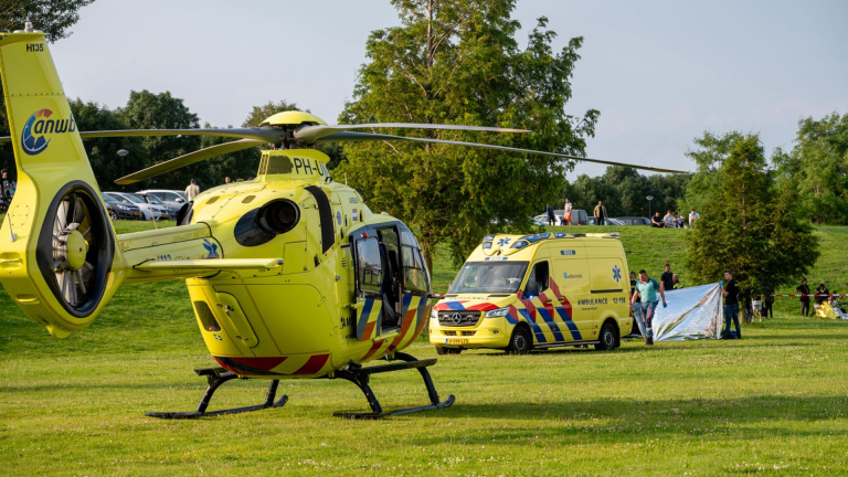 Gele traumahelikopter naast een ambulance op een grasveld met hulpverleners en toeschouwers.