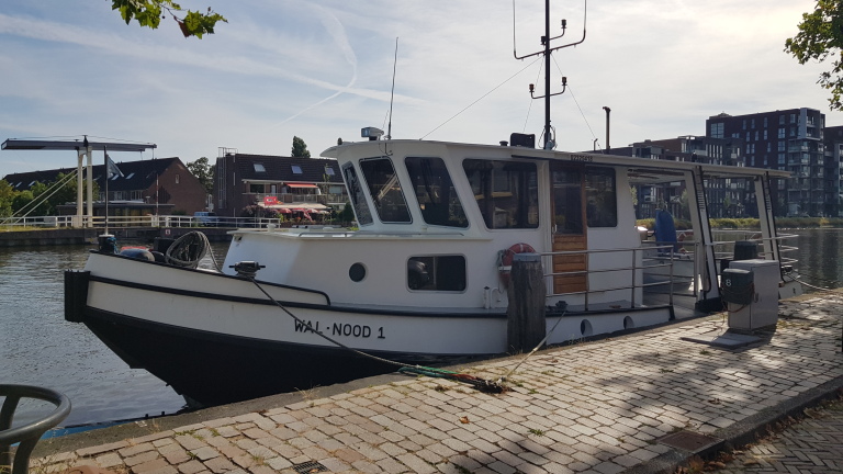 Een witte boot met de naam "WAL NOOD 1" ligt aangemeerd aan een kade naast een kanaal, met huizen en moderne gebouwen op de achtergrond.