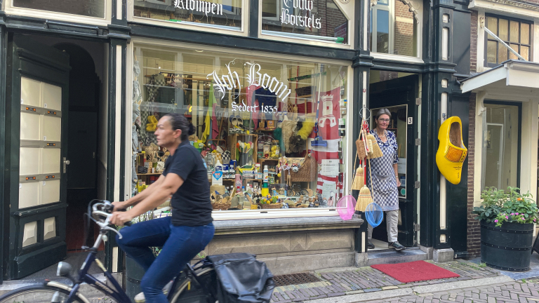 Fietsster rijdt langs een ouderwetse winkel met traditionele producten, met een vrouw in schort in de deuropening en een grote gele klomp aan de gevel.