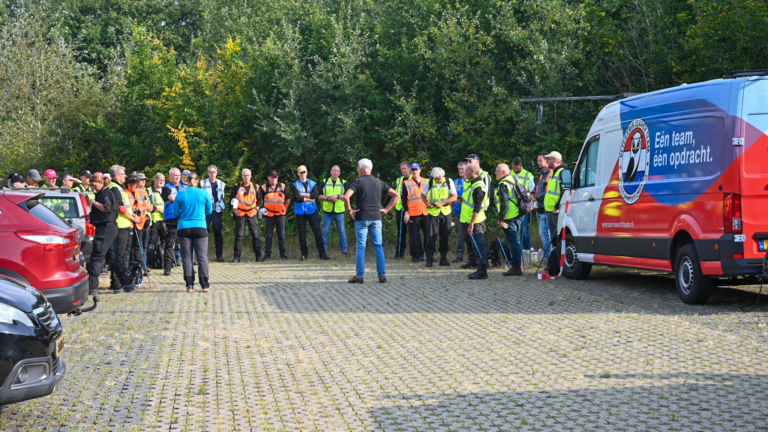 Groep mensen met veiligheidsvesten verzameld op een parkeerterrein bij een witte bestelwagen met de tekst: "Eén team, één opdracht".