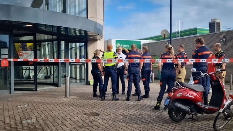 Politie- en brandweerpersoneel staat buiten een afgesloten ingang van een gebouw, met rood-wit afzetlint en een rode scooter op de voorgrond.