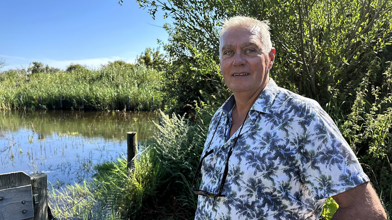 Man in een overhemd met palmboomprint staat naast een rustig beekje en hoge grassen in een natuurlijke omgeving.