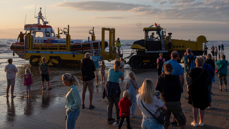 Mensen kijken naar een KNRM-reddingsboot die met een tractor het water in wordt gelanceerd bij zonsondergang.