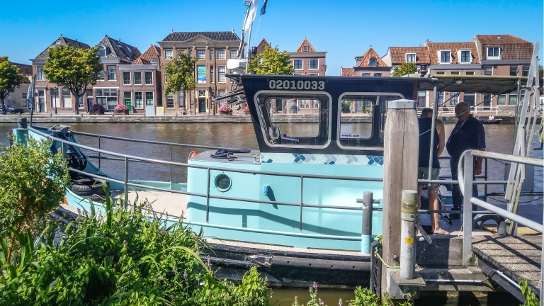 Een lichtblauwe boot aangemeerd aan een steiger, met op de achtergrond grachtenpanden en bomen langs een kanaal op een zonnige dag.