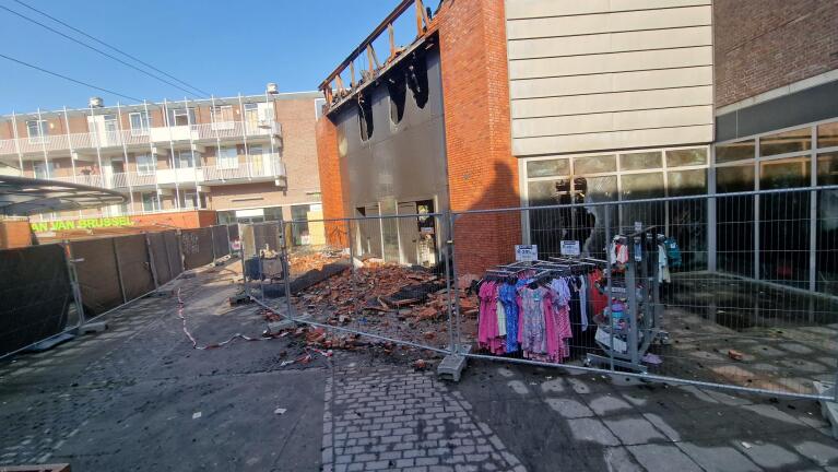 Beschadigd gebouw na een brand, omgeven door hekken en puin, met kledingrekken op de voorgrond.