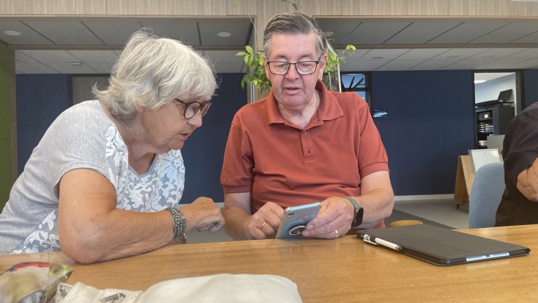 Twee oudere mensen zitten aan tafel en kijken samen naar een smartphone.