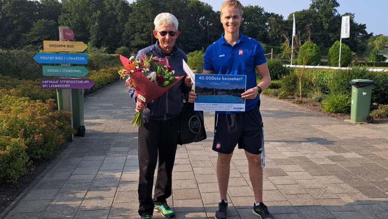 Twee mannen staan buiten, één met een bos bloemen en de ander met een bord waarop "40.000ste bezoeker!" staat. Op de achtergrond zijn gekleurde bewegwijzering en groene struiken te zien.