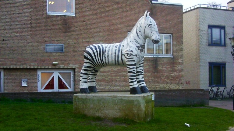 Standbeeld van een beschilderd paard met zwart-witte zebrapatronen op een verhoging voor een bakstenen gebouw.