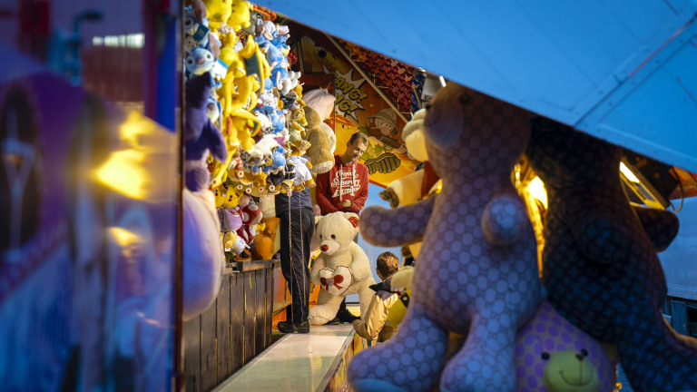 Een kraam op een kermis vol met grote pluchen knuffels en kleurrijke dieren, met een man die een grote teddybeer vasthoudt.