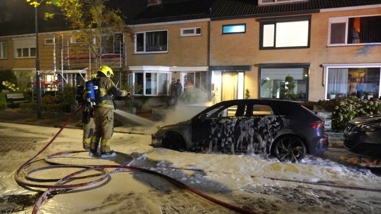 Brandweerman blust brandende auto voor een rijtjeshuis in een woonwijk in de nacht.