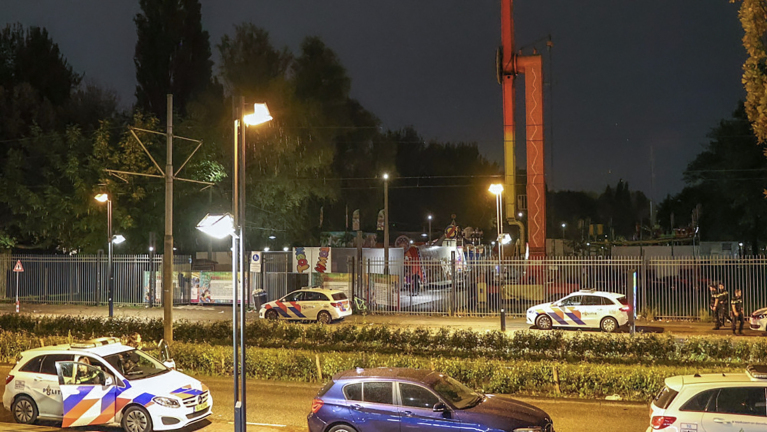 Politieauto's voor een verlicht hek, met een kermisattractie zichtbaar op de achtergrond, 's nachts.