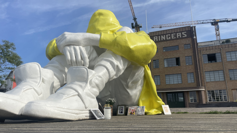 Grote beeldhouwwerk van een figuur in een gele capuchon, zittend met opgetrokken knieën, omringd door foto's en kaarsen, voor een gebouw met de tekst "Ringers".