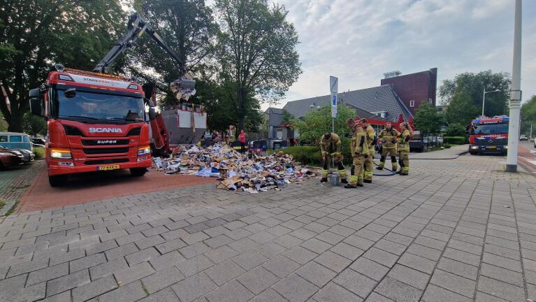 Brandweerlieden blussen een brand in een vuilniswagen, terwijl papierafval op straat ligt, met brandweerwagens op de achtergrond.