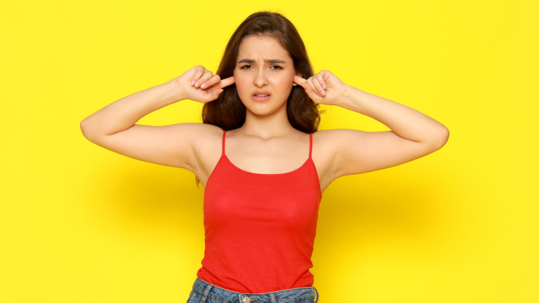 Vrouw met een rode top houdt haar vingers in haar oren tegen een gele achtergrond.