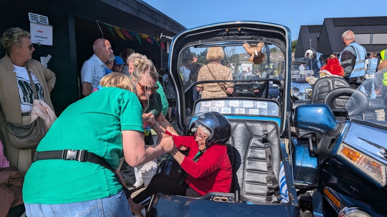 Oudere vrouw met helm zit in een zijspan en krijgt hulp van een medewerker terwijl omstanders toekijken.