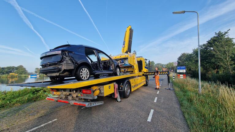 Autowrak wordt op een bergingswagen geladen naast een weg met een waterlichaam en een verkeersbord met de tekst "West-Graftdijk".