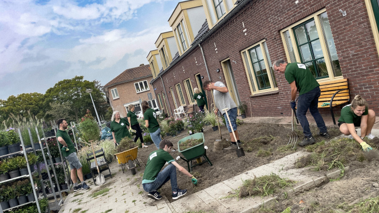 Groep mensen in groene shirts werken gezamenlijk in een tuin voor een gebouw met bakstenen muren en grote ramen.