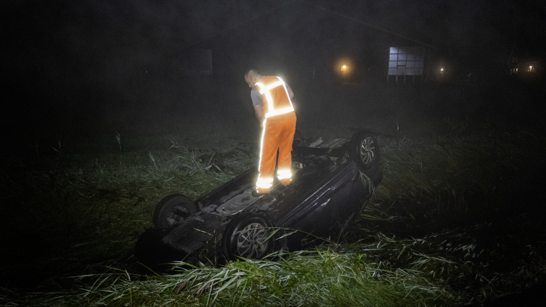 Man in reflecterend veiligheidsvest staat bovenop een omgeslagen auto in een grasveld in het donker.