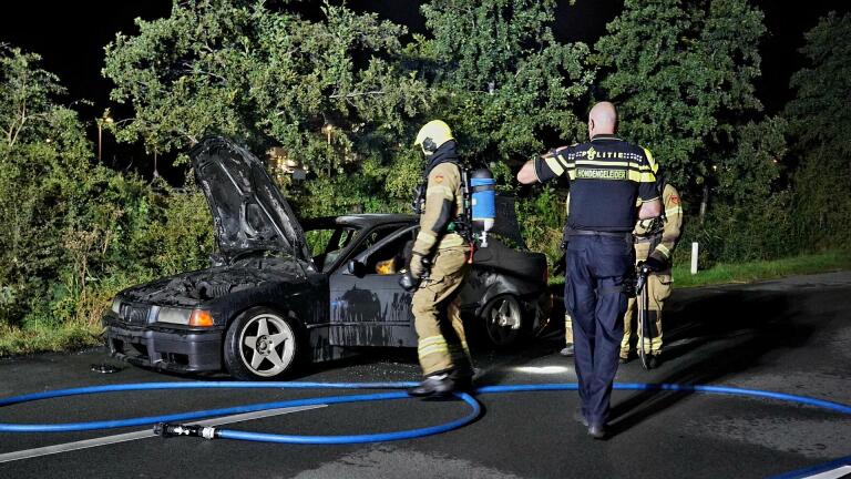 Brandweerlieden en een politieagent onderzoeken een uitgebrande auto langs de weg in de nacht.