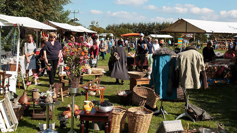 Mensen die in de zon rondlopen en winkelen op een drukke buitenmarkt met verschillende kraampjes en producten.