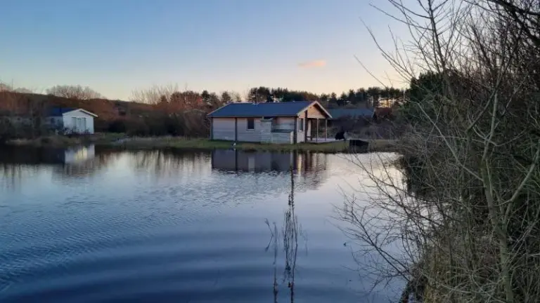 Houten huis aan de rand van een meer bij zonsondergang met een bos in de achtergrond.