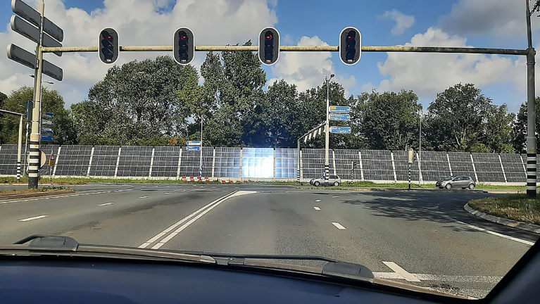 Kruispunt met verkeerslichten gezien vanuit een auto, met zonnecollectoren en bomen op de achtergrond.