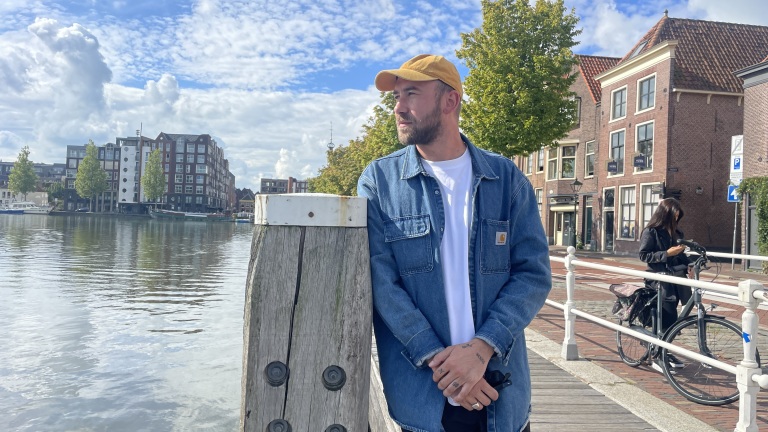 Man leunend tegen een houten paal aan een gracht, met gebouwen en een vrouw met een fiets op de achtergrond.