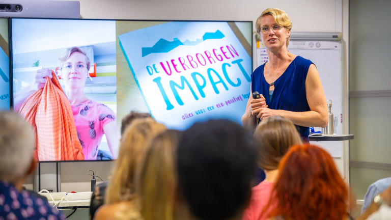 Vrouw geeft een presentatie voor een groep mensen met de titel "De Verborgen Impact" op het scherm achter haar.