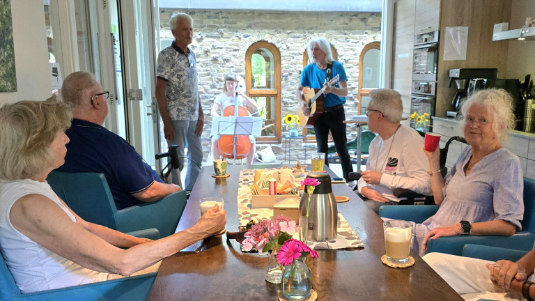 Mensen zitten aan een tafel in een keuken terwijl muzikanten op een cello en gitaar spelen.