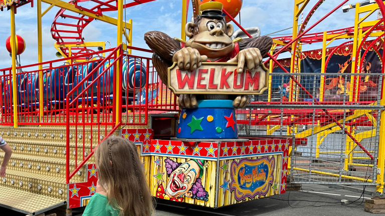 Grote opgezette aap die een bord met "Welkom" vasthoudt bij de ingang van een kleurrijke kermisattractie.