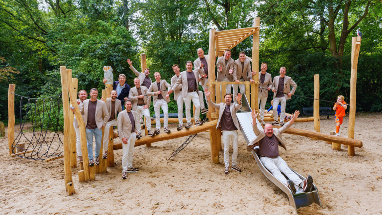 Een groep mannen in beige en bruine kleding poseert op een houten speeltuin in een bosrijke omgeving. Een kind in een oranje outfit speelt op de achtergrond.