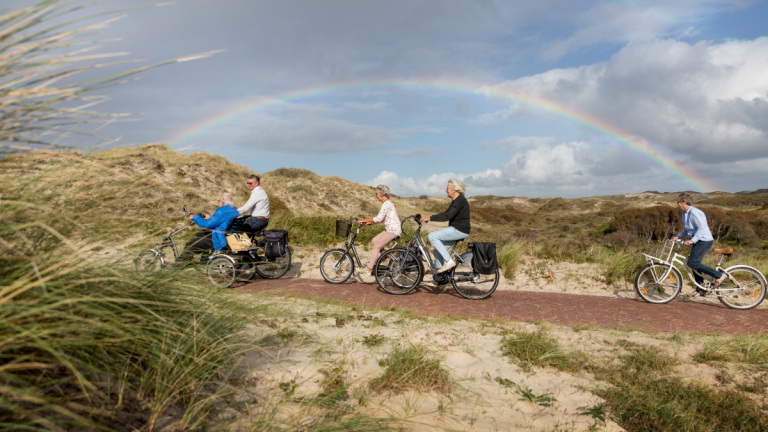 Mensen fietsen over een pad door de duinen met een regenboog in de lucht.