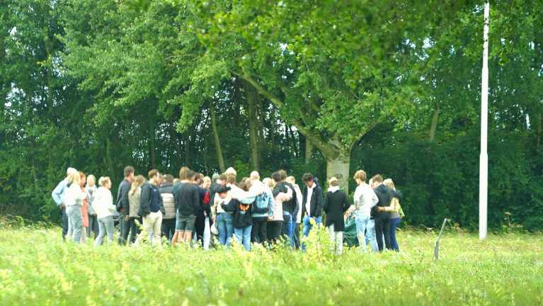 Groep mensen verzameld in een open veld met bomen op de achtergrond.