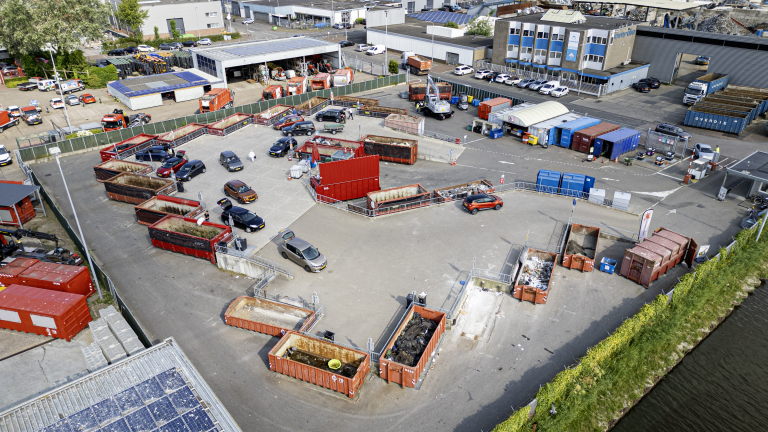 Luchtfoto van een milieupark met meerdere containers en voertuigen die afval weggooien. Op de achtergrond zijn bedrijfsgebouwen en een parkeerplaats te zien.