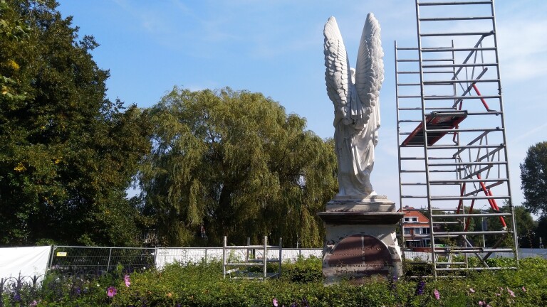 Standbeeld van een engel naast een bouwsteiger, omringd door een tuin met bloemen en bomen op de achtergrond.