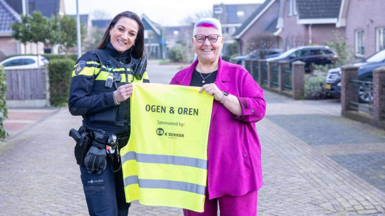 Politieagent en oudere vrouw met roze haren houden gezamenlijk een veiligheidsvest vast met de tekst "OGEN & OREN" op een rustige straat.