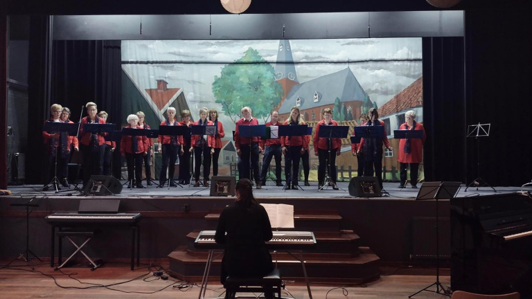 Een koor in rode kleding zingt op een toneel met een landschap-decor op de achtergrond, terwijl een vrouw piano speelt op de voorgrond.