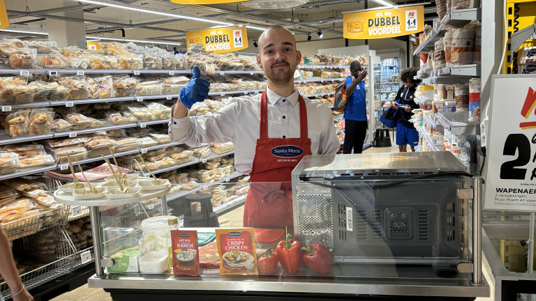 Man in een supermarkt met een rode schort van Santa Maria geeft een duim omhoog naast een proeverijtafel met dips en ingrediënten.