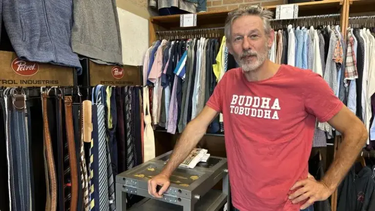 Man in kledingwinkel met rode 'Buddha-to-Buddha' T-shirt, omringd door kleding en accessoires.