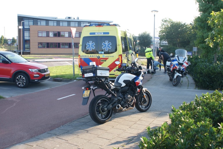 Ambulance en politie bij een incident op straat met een politieagent die iemand onderzoekt, naast een gele ambulance en twee politie motorfietsen.