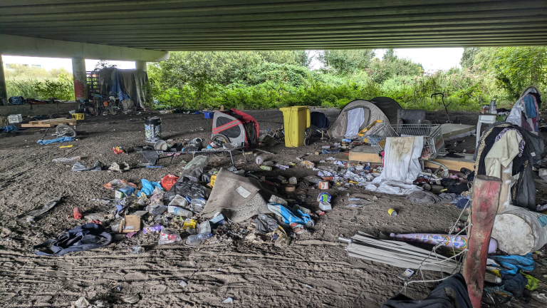 Kamp van daklozen onder een viaduct met tenten, fietsen en verspreid afval.