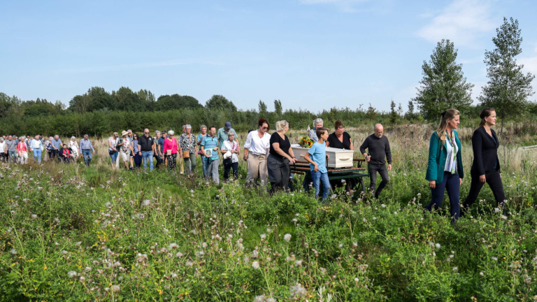 Een groep mensen in een groene, natuurlijke omgeving voert een uitvaartstoet aan, waarbij een kist op een kar geduwd wordt.