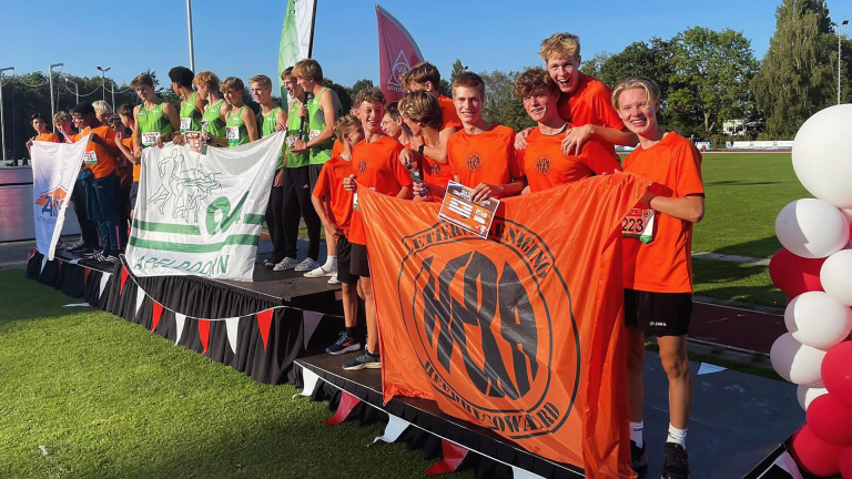 Atletiekteams poseren op het podium met hun vlaggen tijdens een sportevenement op een zonnige dag.