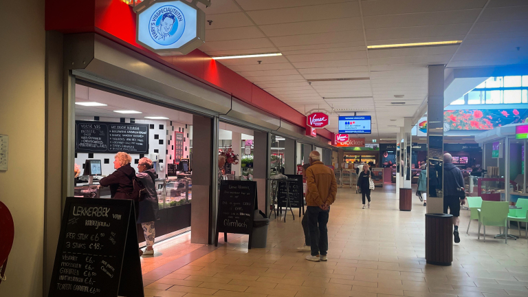 Gangpad in een winkelcentrum met verschillende winkels, waaronder een viswinkel genaamd "Perry's Visspeciaaliteiten" waar klanten worden bediend, en een Vomar supermarkt op de achtergrond.