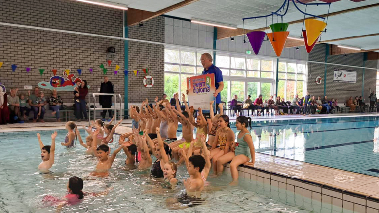 Kinderen vieren het behalen van hun zwemdiploma in een binnenzwembad, terwijl een instructeur een diploma omhoog houdt.