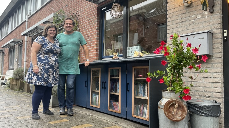 Man en vrouw staan voor hun huis naast een blauwe buurtbibliotheek met veel boeken, planten en een postbus.