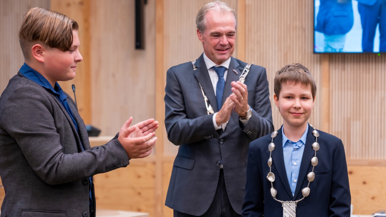 Drie personen in formele kleding, waarvan twee volwassenen en een kind, staan in een zaal. Het kind draagt een ambtsketen en de volwassenen klappen.