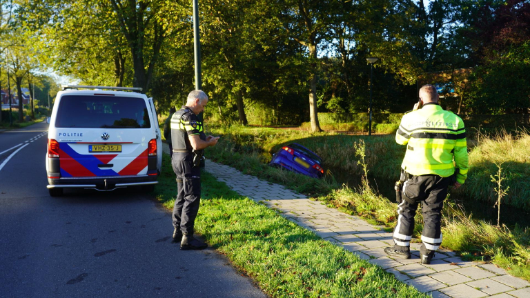 Twee politieagenten naast een politiebusje onderzoeken een blauwe auto die in een sloot is gereden.