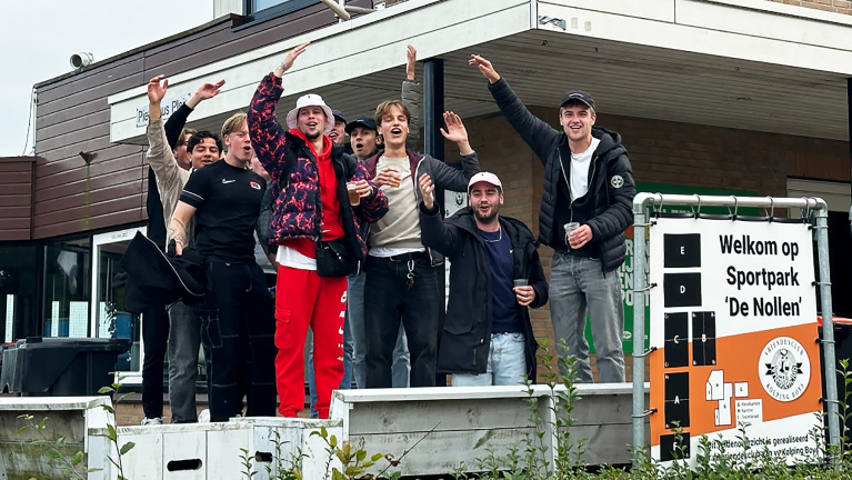 Groep jonge mannen juichen en poseren voor de ingang van Sportpark 'De Nollen' met een bord op de voorgrond.